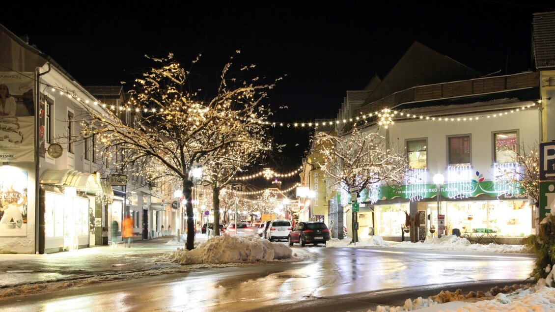 Eine schneebedeckte Straße in der Stadt bei Nacht, erleuchtet von festlichen Lichtern. Weiße Bäume und geschmückte Geschäfte schaffen eine gemütliche Atmosphäre. | © thumbnail_Advent in der Stadt_Hauptplatz Foto Sima Susi