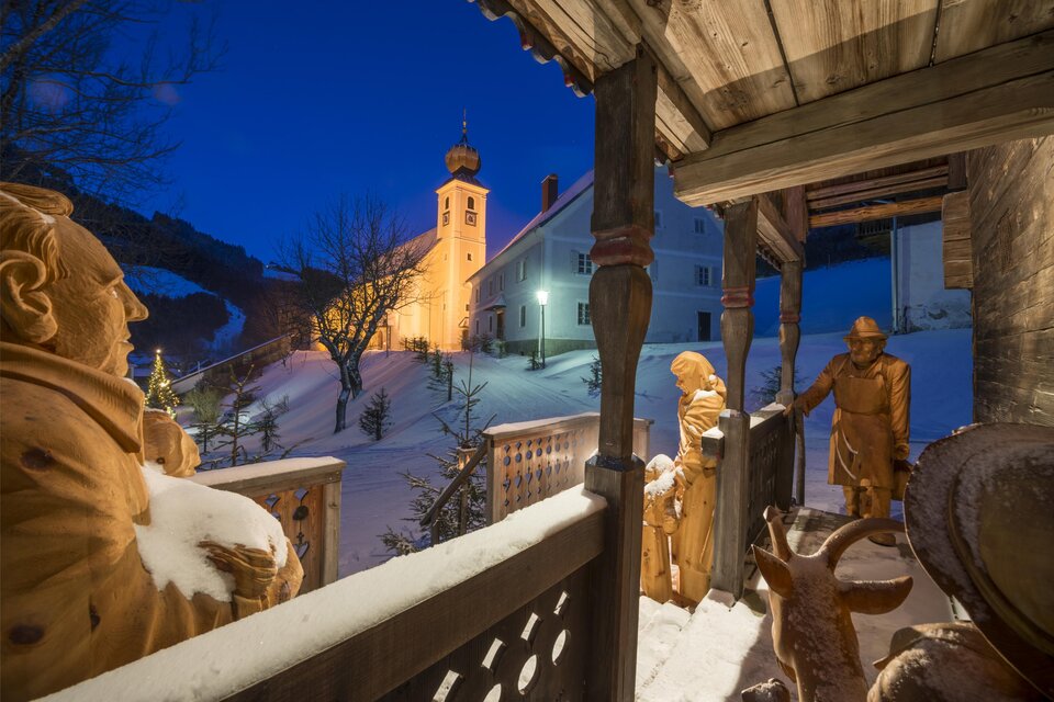 Eine verschneite Winterlandschaft mit einer beleuchteten Kirche. Skulpturen und Menschen stehen auf einer überdachten Terrasse. | © Tourismusverband Oststeiermark