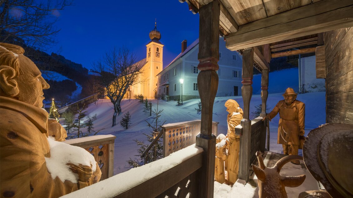 Eine verschneite Winterlandschaft mit einer beleuchteten Kirche. Skulpturen und Menschen stehen auf einer überdachten Terrasse. | © Tourismusverband Oststeiermark