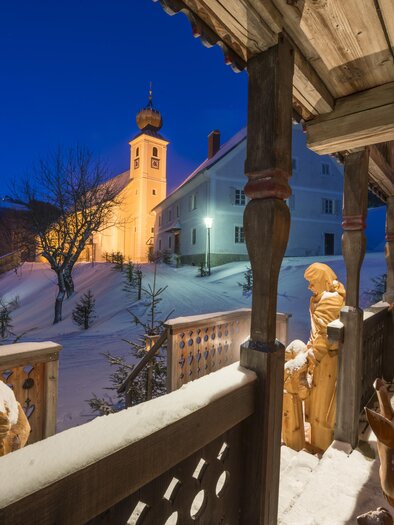 Egy havas téli táj egy kivilágított templommal. Szobrok és emberek állnak egy fedett teraszon. | © Tourismusverband Oststeiermark