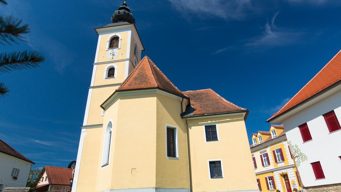 Eine schöne Kirche mit einem hohen Turm und einem orangefarbenen Dach. Der Himmel ist klar und blau, umgeben von bunten Gebäuden. | © Region Graz-Harry Schiffer