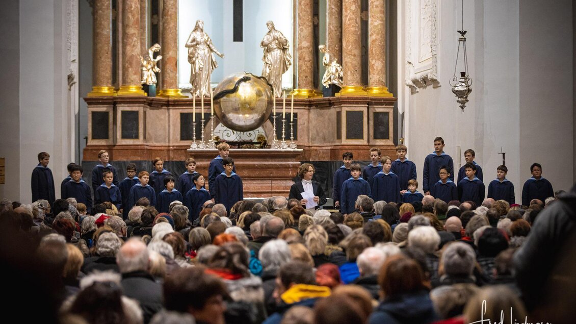 Ein Kirchenkonzert mit einem Kinderchor auf der Bühne vor einer großen Menschenmenge. Die Atmosphäre ist festlich und der Altar ist dekorativ gestaltet. | © Fred Lindmoser