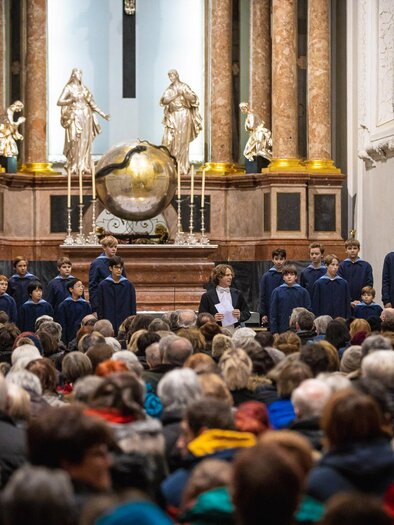 A church concert with a children's choir on stage in front of a large crowd. The atmosphere is festive and the altar is decoratively designed. | © Fred Lindmoser