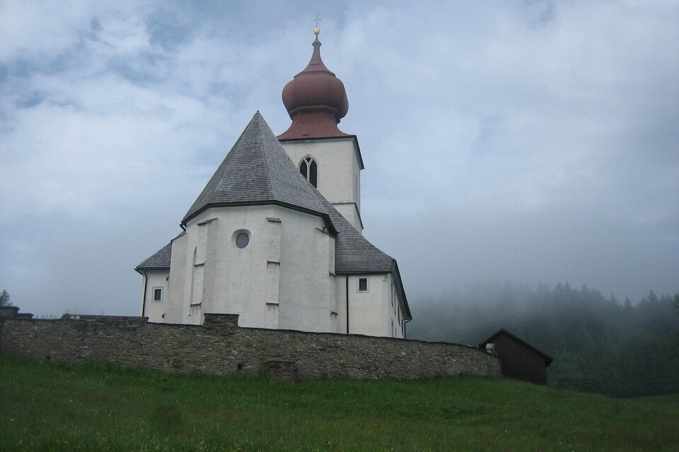 Eine kleine Kirche mit einer markanten roten Kuppel steht in einer bewachsenen Wiese. Im Hintergrund sind Nebel und Bäume sichtbar.