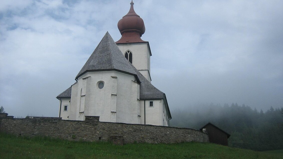 Eine kleine Kirche mit einer markanten roten Kuppel steht in einer bewachsenen Wiese. Im Hintergrund sind Nebel und Bäume sichtbar.