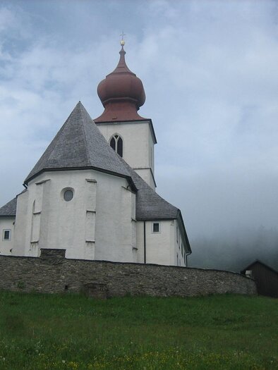 Eine kleine Kirche mit einer markanten roten Kuppel steht in einer bewachsenen Wiese. Im Hintergrund sind Nebel und Bäume sichtbar.