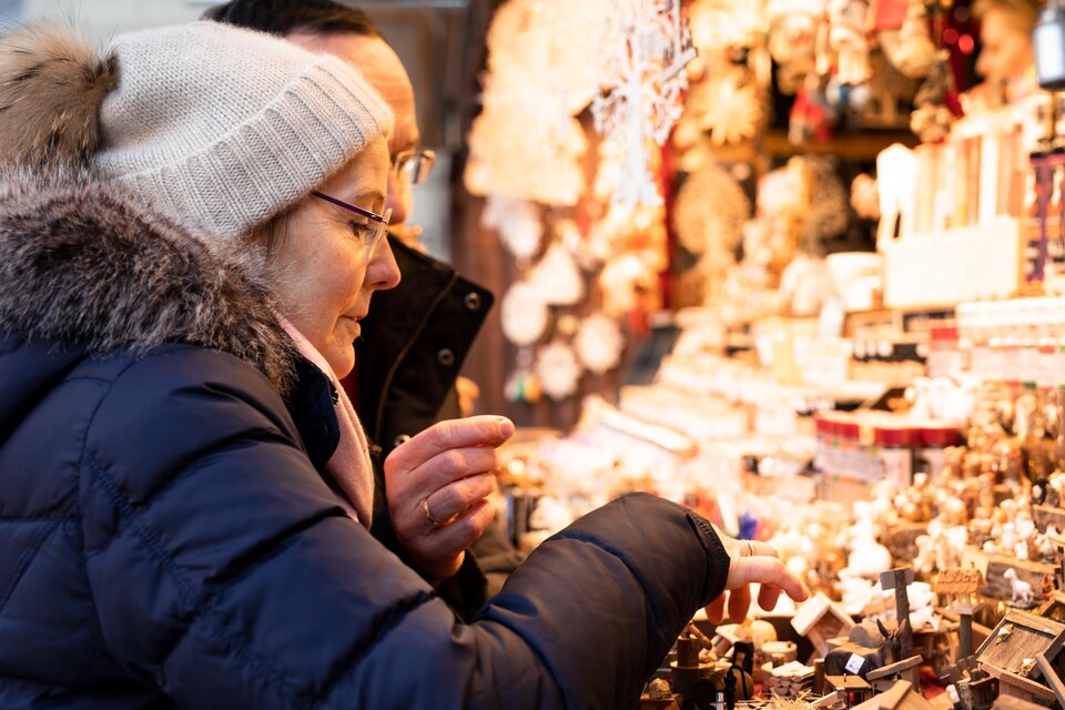 Eine Frau betrachtet sorgfältig kleine Holzfiguren auf einem Marktstand. Im Hintergrund sind weitere festliche Dekorationen sichtbar. | © Graz Tourismus