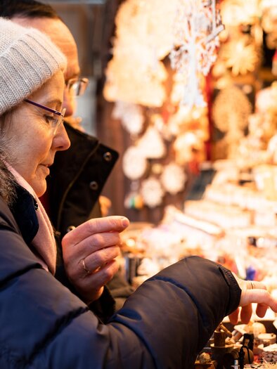 A woman carefully examines small wooden figures at a market stall. In the background, more festive decorations are visible. | © Graz Tourismus