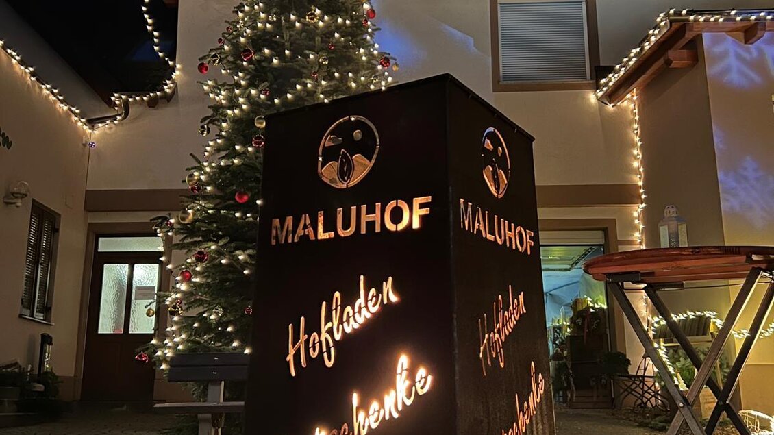 Ein schön dekorierter Innenhof mit einem Weihnachtsbaum und Lichtern. Im Vordergrund steht ein beleuchtetes Schild mit Informationen zum Maluhof. | © MALU