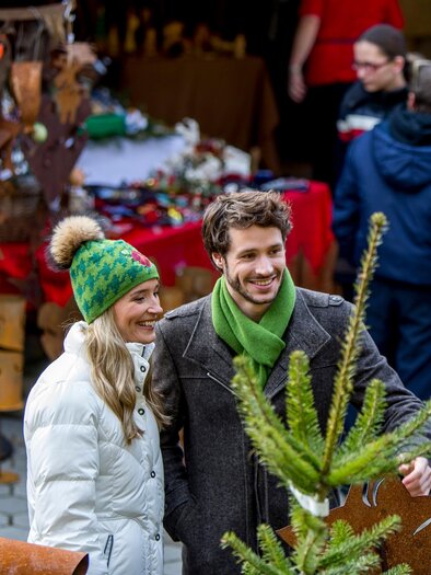 Ein Paar lächelt in einem festlichen Weihnachtsmarkt, umgeben von handgemachten Ornamenten und Dekorationen. Im Hintergrund sind weitere Besucher zu sehen, die den Markt erkunden. | © Region Graz-Tom Lamm