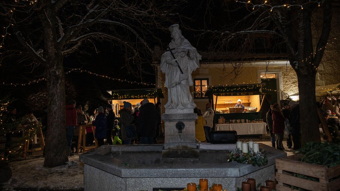 Eine festlich dekorierte Szene mit einer Statue im Zentrum. Im Hintergrund sind zahlreiche Menschen und Lichter zu sehen, die eine gemütliche Atmosphäre schaffen. | © Heinz&Melitta Holzmüller