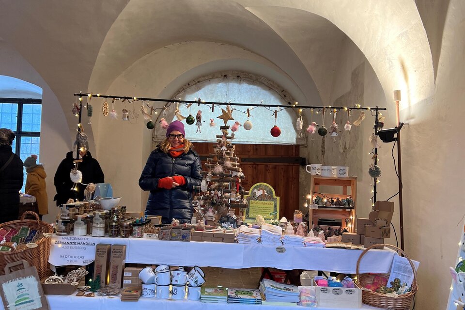 Ein Weihnachtsmarktstand in einem historisch anmutenden Raum. Der Stand ist dekoriert mit festlichen Produkten und Lichtern. | © Lisa-Marie Schiefer