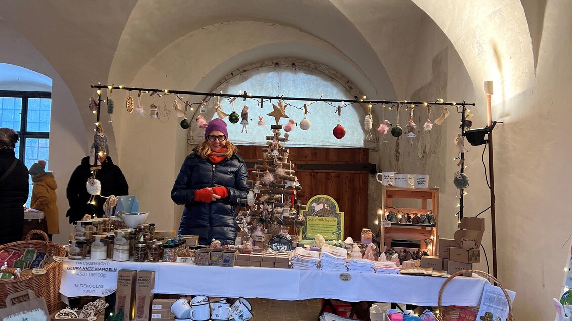 Ein Weihnachtsmarktstand in einem historisch anmutenden Raum. Der Stand ist dekoriert mit festlichen Produkten und Lichtern. | © Lisa-Marie Schiefer