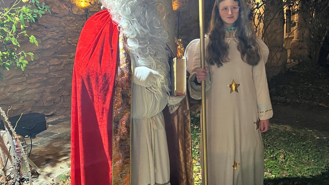 Ein traditioneller Nikolaus steht mit einem Engel in einem Garten. Der Nikolaus trägt einen roten Mantel und hält einen goldenen Stab, während das Engelkind einen Korb mit Geschenken bei sich hat. | © Lisa-Marie Schiefer