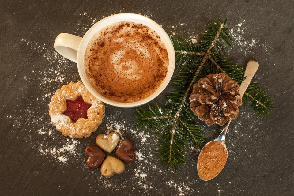 Eine Tasse heiße Schokolade mit Sahne und Zimt, umgeben von Weihnachtsgebäck und Tannenzweigen. Im Hintergrund sind etwas Puderzucker und eine Kieferzapfen zu sehen. | © Tourismusverband Schladming-Dachstein