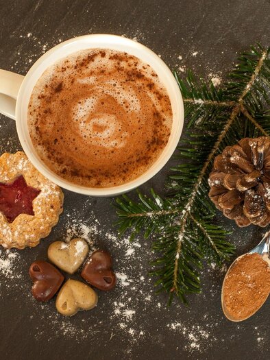 Eine Tasse heiße Schokolade mit Sahne und Zimt, umgeben von Weihnachtsgebäck und Tannenzweigen. Im Hintergrund sind etwas Puderzucker und eine Kieferzapfen zu sehen. | © Tourismusverband Schladming-Dachstein