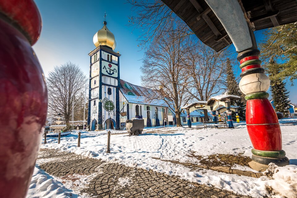 Eine bunte Kirche mit goldener Kuppel steht in einer verschneiten Landschaft. Dahinter sind einige Bäume und ein klarer blauer Himmel sichtbar. | © Lipizzanerheimat-Die Abbilderei