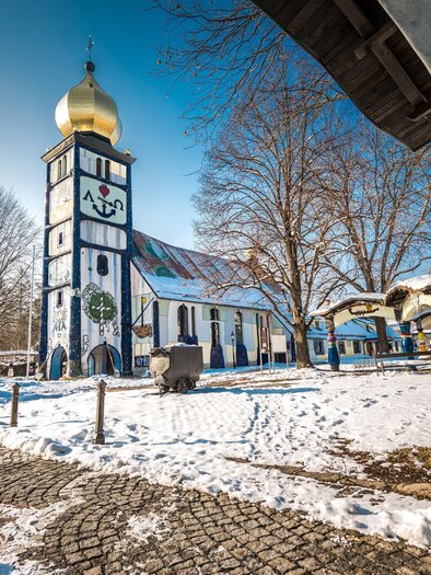 Eine bunte Kirche mit goldener Kuppel steht in einer verschneiten Landschaft. Dahinter sind einige Bäume und ein klarer blauer Himmel sichtbar. | © Lipizzanerheimat-Die Abbilderei