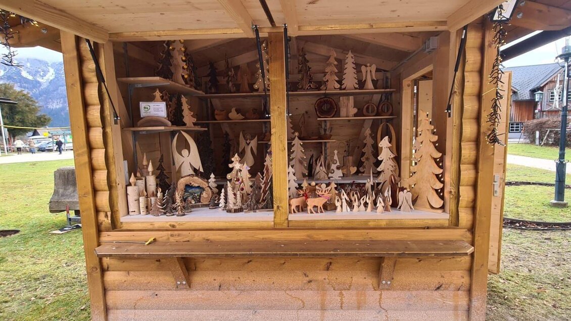 Ein Holzstand mit feiner handgefertigter Dekoration, darunter viele kleine Holzfiguren und Bäume. Der Stand befindet sich in einer malerischen Umgebung mit Bergen im Hintergrund. | © Winfried Büttner
