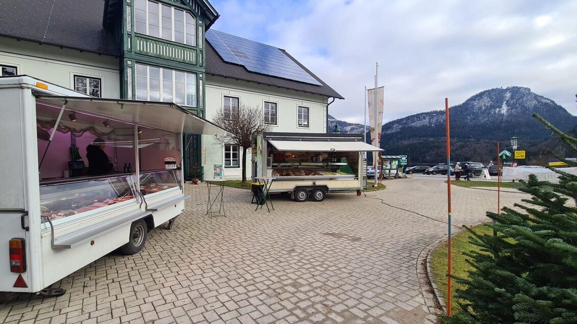 Ein Markt mit zwei Verkaufsanhängern steht vor einem großen Gebäude. Im Hintergrund sind Berge und ein bewölkter Himmel zu sehen. | © Winfried Büttner