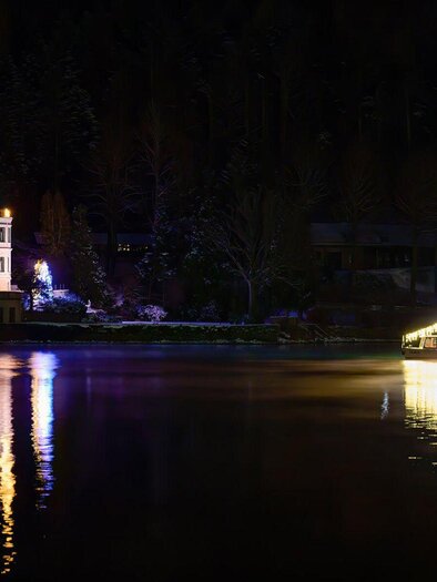 Die malerische Villa Castiglioni am Ufer des ruhigen Grundlsees, umgeben von verschneiten Bäumen. Im Wasser des Grundlsees spiegelt sich das Licht eines beleuchteten Bootes – der MS Traun von der Schifffahrt Grundlsee. | © Schifffahrt Grundlsee