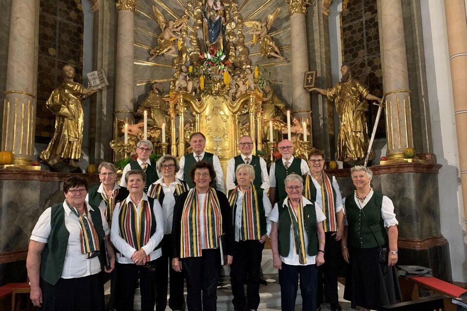 Eine Gruppe von Sängerinnen und Sängern steht vor einem prächtigen Altar in einer Kirche. Alle tragen festliche Kleidung und Schal mit Streifen. | © Vulkanlandchor Pertlstein