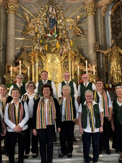 Eine Gruppe von Sängerinnen und Sängern steht vor einem prächtigen Altar in einer Kirche. Alle tragen festliche Kleidung und Schal mit Streifen. | © Vulkanlandchor Pertlstein
