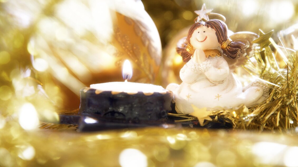 Eine kleine Figur eines Engels sitzt neben einer brennenden Kerze. Im Hintergrund funkeln goldene Lichter, die eine festliche Stimmung erzeugen. | © Steiermark Tourismus-Photostyle.at