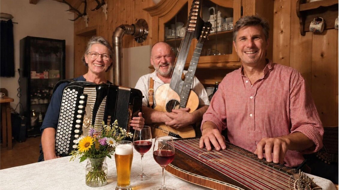 <p>Drei Musiker der Gruppe Wallberg sitzen an einem Tisch und spielen traditionelle Instrumente. Auf dem Tisch stehen Getränke und eine Glasvase mit Wiesenblumen.</p> | © Wallberg Trio