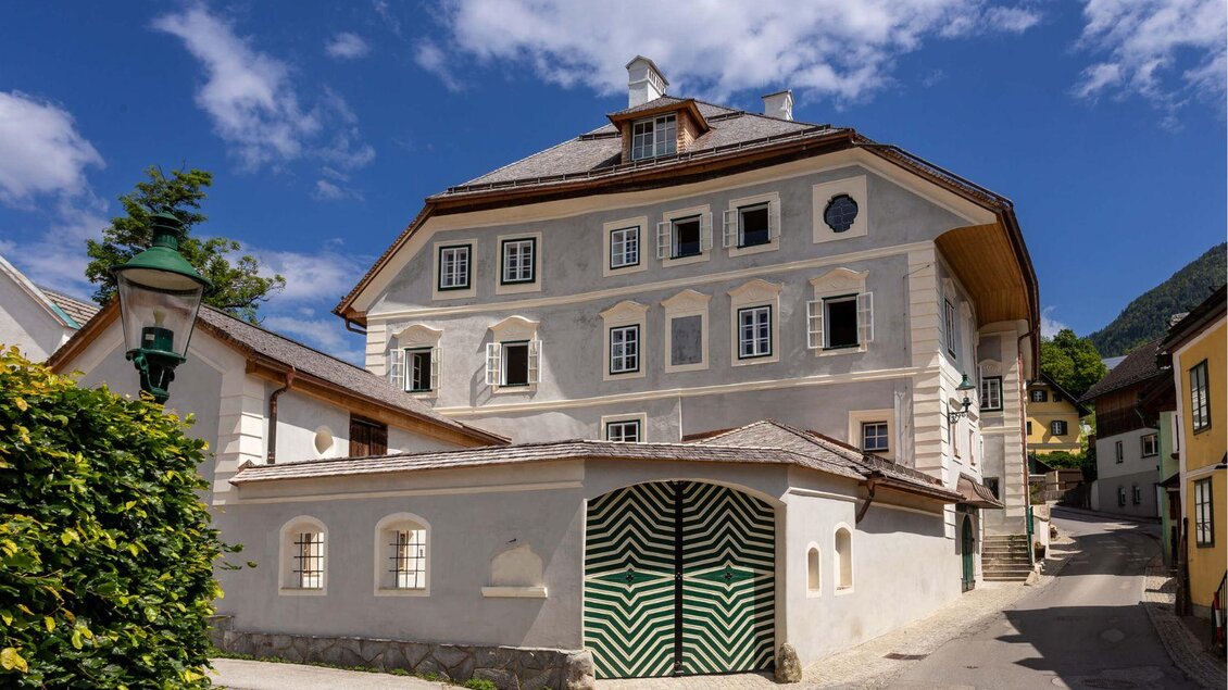 Das historische Gebäude des Podenhaus mit charakteristischer Fassade und grunem Tor mit weißen Details. Die Straße der Kirchengasse in Bad Aussee ist ruhig und von Bäumen gesaumt. | © Podenhaus, Bad Aussee