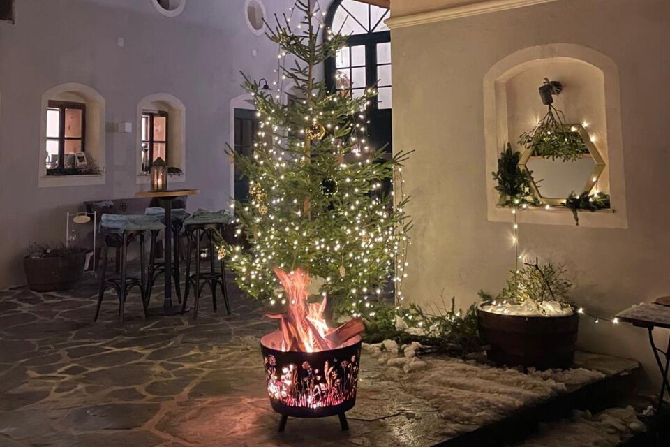 Ein festlich dekorierter Weihnachtsbaum mit Lichtern steht in einem gemütlichen Raum. Im Vordergrund brennt ein Lagerfeuer in einem runden Feuerkorb. | © Podenhaus, Bad Aussee