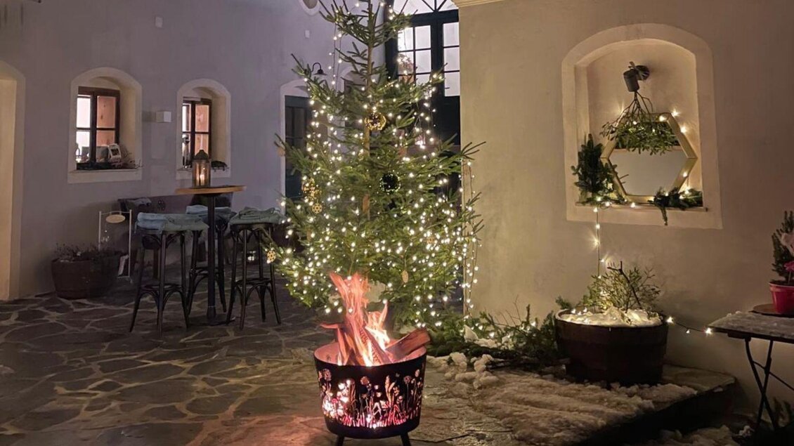 Ein festlich dekorierter Weihnachtsbaum mit Lichtern steht in einem gemütlichen Raum. Im Vordergrund brennt ein Lagerfeuer in einem runden Feuerkorb. | © Podenhaus, Bad Aussee