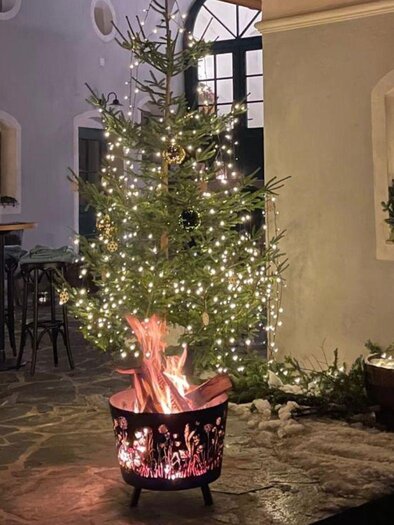 Ein festlich dekorierter Weihnachtsbaum mit Lichtern steht in einem gemütlichen Raum. Im Vordergrund brennt ein Lagerfeuer in einem runden Feuerkorb. | © Podenhaus, Bad Aussee