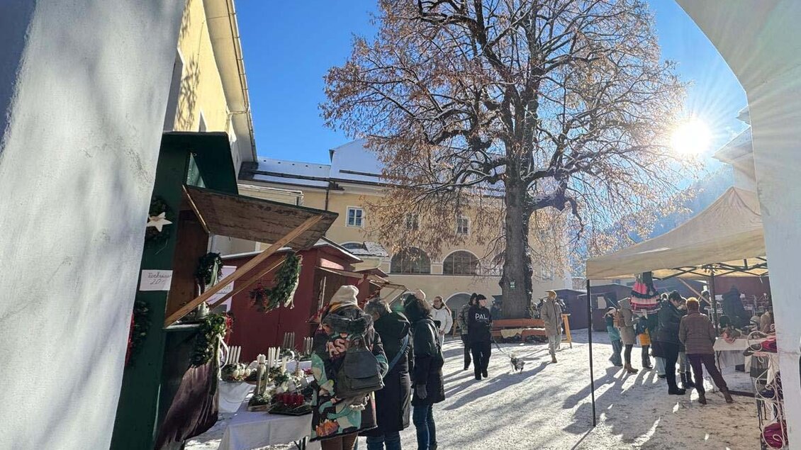 Ein lebhafter Weihnachtsmarkt mit Menschen, die lokale Produkte kaufen. Im Hintergrund steht ein großer Baum und die Sonne scheint strahlend auf die Szene. | © Elternverein Donnersbach