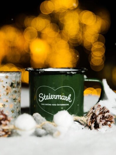 A green mug next to a sparkling candle holder sits on a snow-covered surface. In the background, soft lights twinkle, and there are some pine cones.