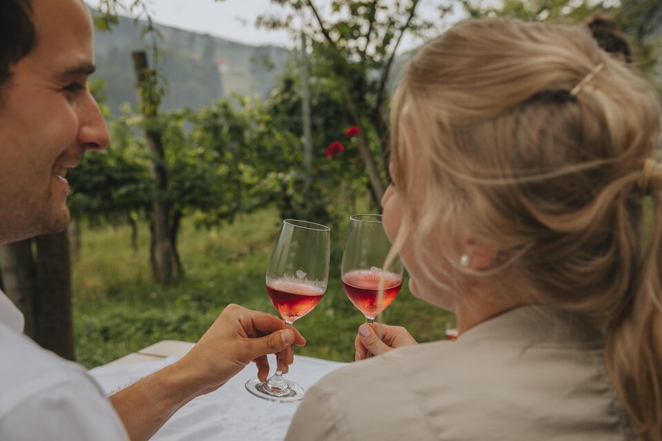 Ein Paar stößt mit Gläsern auf einem Weinberg an. Im Hintergrund sind grüne Landschaften zu sehen. | © Lupi Spuma