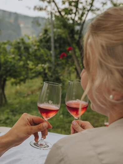 Ein Paar stößt mit Gläsern auf einem Weinberg an. Im Hintergrund sind grüne Landschaften zu sehen. | © Lupi Spuma