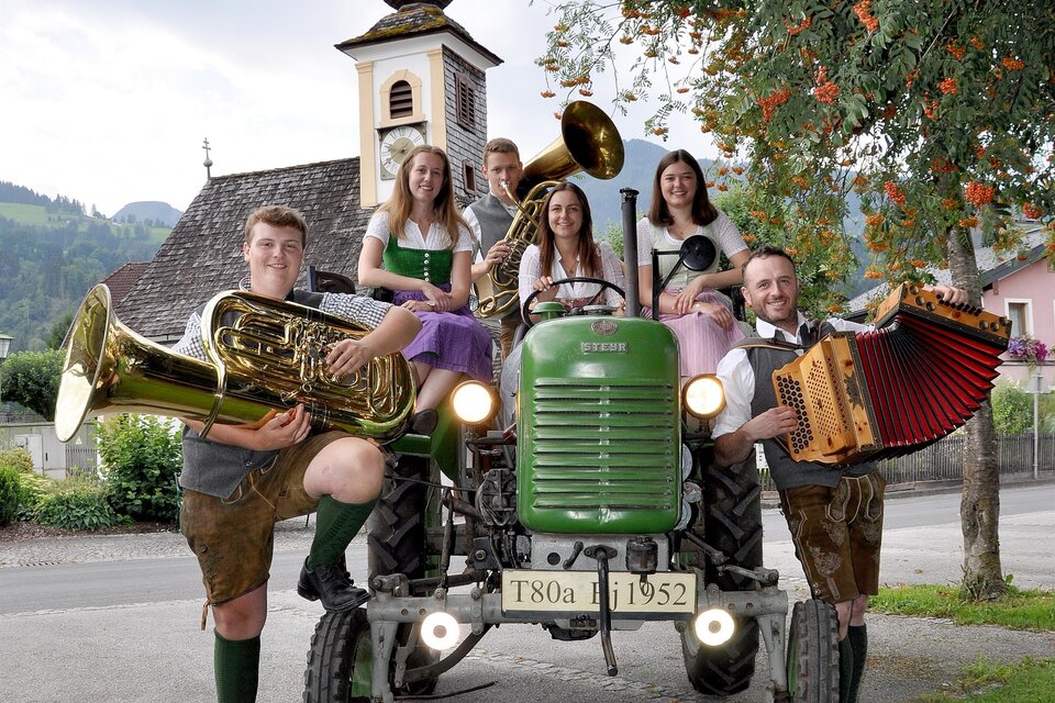 Eine Gruppe von Menschen in traditioneller Kleidung steht um einen alten Traktor. Sie halten Musikinstrumente und posieren vor einer malerischen Kulisse mit einem Kirchturm im Hintergrund. | © IDA/ARGE Aicher Kirtag