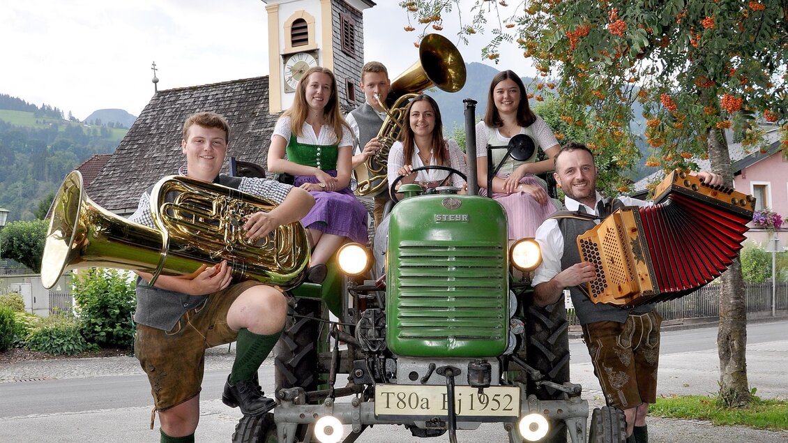 Eine Gruppe von Menschen in traditioneller Kleidung steht um einen alten Traktor. Sie halten Musikinstrumente und posieren vor einer malerischen Kulisse mit einem Kirchturm im Hintergrund. | © IDA/ARGE Aicher Kirtag