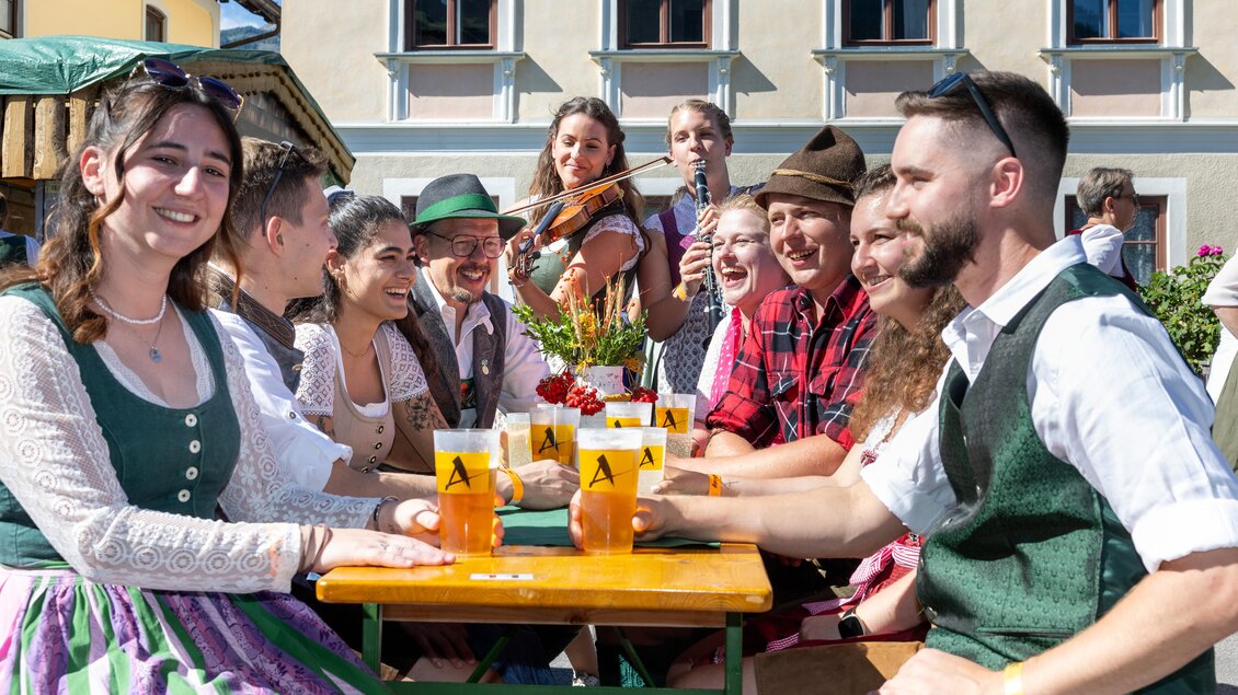 Eine fröhliche Gruppe von Menschen sitzt an einem Tisch und genießt Getränke. Im Hintergrund spielt eine Musikerin auf der Geige, während alle miteinander lachen und sich amüsieren. | © ARGE Aicher Kirtag©Harald Steiner