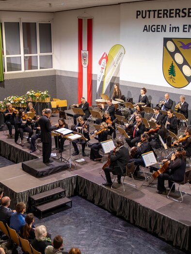 A music teacher conducts an orchestra in the Putterssehalle. The audience sits captivated and enjoys the performance. | © Gemeinde Aigen im Ennstal