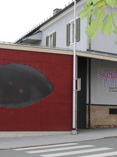 Ein Gebäude mit einem großen, künstlerischen Augemotiv an der Wand. Die Architektur ist modern und die Umgebung ist ruhig. | © TV Südsteiermark - Irene Löschnig