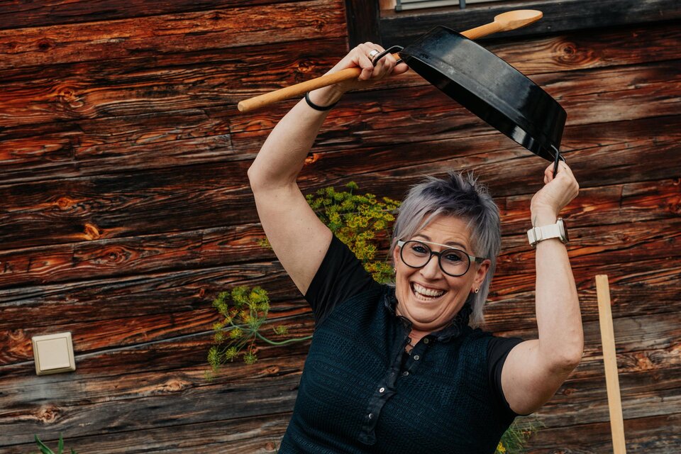 Eine fröhliche Frau steht vor einer Holzwand und hebt eine Pfanne in die Luft. Sie trägt eine schwarze Bluse und eine kurze Hose und scheint sich zu amüsieren. | © studio draussen