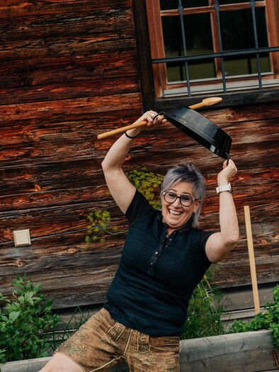 Eine fröhliche Frau steht vor einem Holzhaus und hält eine Pfanne über ihrem Kopf. Sie trägt ein schwarzes Shirt und Shorts und hat graue Haare. | © studio draussen