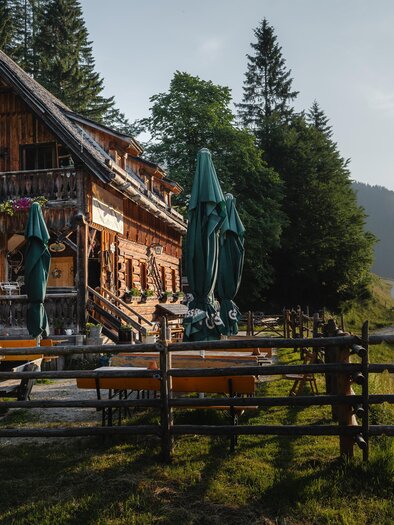 A traditional mountain cabin with a wooden facade stands amidst trees. In the background, gentle hills and mountains are visible, while a gravel path runs by.