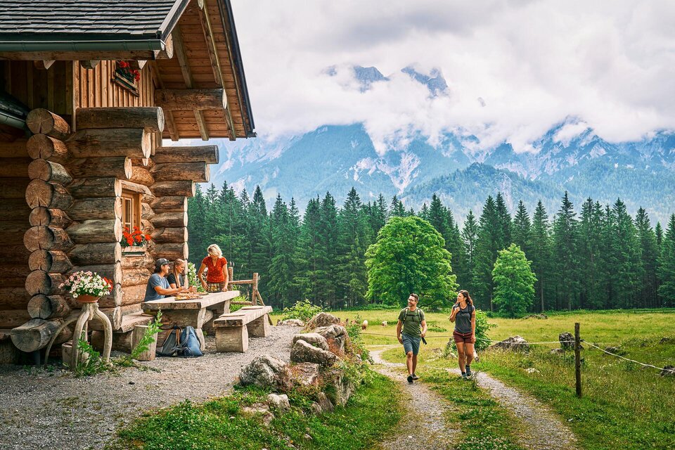 Eine gemütliche Hütte in den Bergen mit einer schönen Umgebung. Zwei Wanderer gehen den Weg entlang, umgeben von grünen Wiesen und Wäldern.