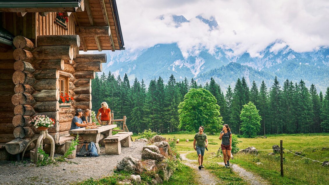 Eine gemütliche Hütte in den Bergen mit einer schönen Umgebung. Zwei Wanderer gehen den Weg entlang, umgeben von grünen Wiesen und Wäldern.