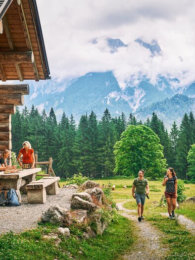 Eine gemütliche Hütte in den Bergen mit einer schönen Umgebung. Zwei Wanderer gehen den Weg entlang, umgeben von grünen Wiesen und Wäldern.