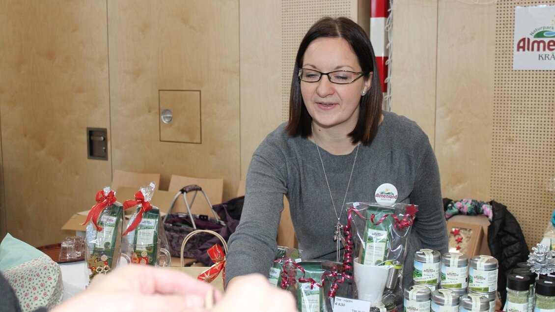 Eine Frau steht an einem Stand mit verschiedenen Produkten, darunter Gläser und Geschenkkörbe. Sie lächelt, während sie mit einem Kunden interagiert. | © Gemeinde Gasen