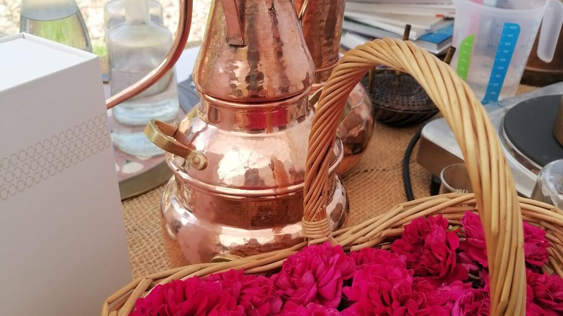 Eine kupferne Kaffeekanne steht neben einem Korb mit leuchtend pinken Blumen. Im Hintergrund sind verschiedene Utensilien und Flaschen sichtbar. | © Sabrina Lukas